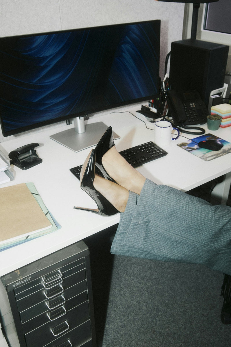 Black heels and dress pants resting on a desk with monitor, keyboard, and notes. CONTRA CLASS — Opening Entry, sensory conscious workwear.